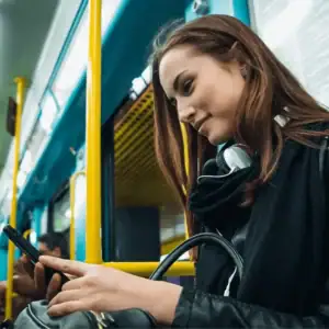 Title: "Top talent engagement on the move"
Description:
Take an authentic snapshot of the modern job seeker. A young professional with straight, long brown hair sits comfortably on public transportation. With fashionable headphones around her neck and a sincere smile, she is captivated by her smartphone - probably exploring trending social media recruiting posts. While keeping a firm grip on her bag in this vibrant commuter environment, highlighted by yellow handrails and blue interior panels, she epitomizes today's dynamic candidate driven by tech-savvy recruitment strategies.
Optimize your recruiting efforts to meet talent where they are! #TalentEngagement #SocialRecruiting #ModernJobSeeker